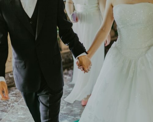 A romantic moment capturing a bride and groom holding hands in elegant wedding attire indoors.